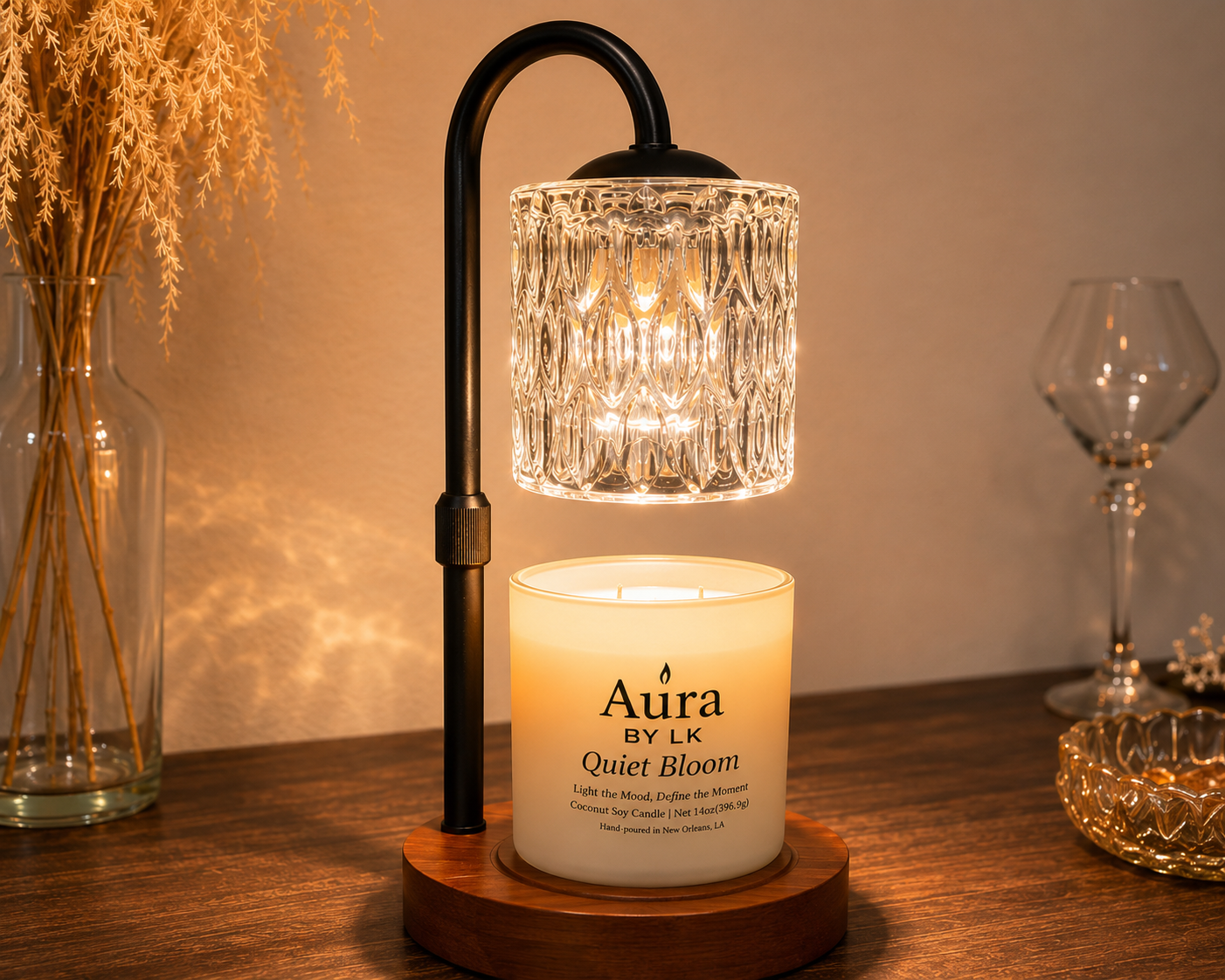A lit scented candle in a white jar labeled 'Aura BY LK Quiet Bloom' sits on a wooden base beneath a black metal arc lamp with a faceted clear glass shade, styled on a wooden table with a wine glass and dried grass in the background.